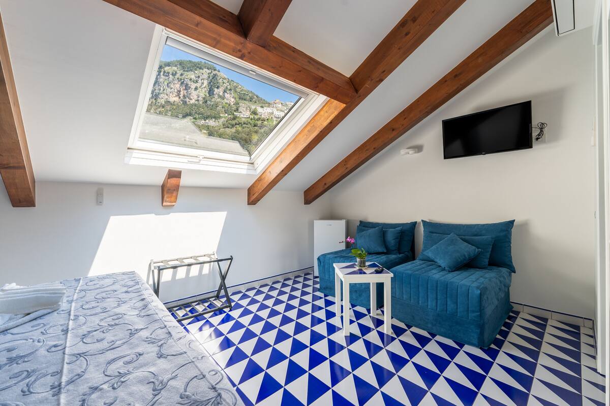 Ein Zimmer mit blau-weiß kariertem Boden, einem blauen Sofa, einem kleinen weißen Tisch, einem an der Wand befestigten Fernseher und einem Fenster mit Blick auf eine Berglandschaft.