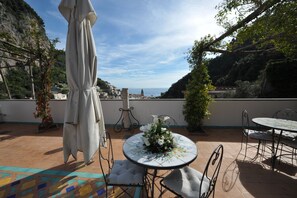 Eine Terrasse im Freien mit einem runden Tisch und Stühlen, einem Sonnenschirm und Blick auf das Meer und die Hügel.