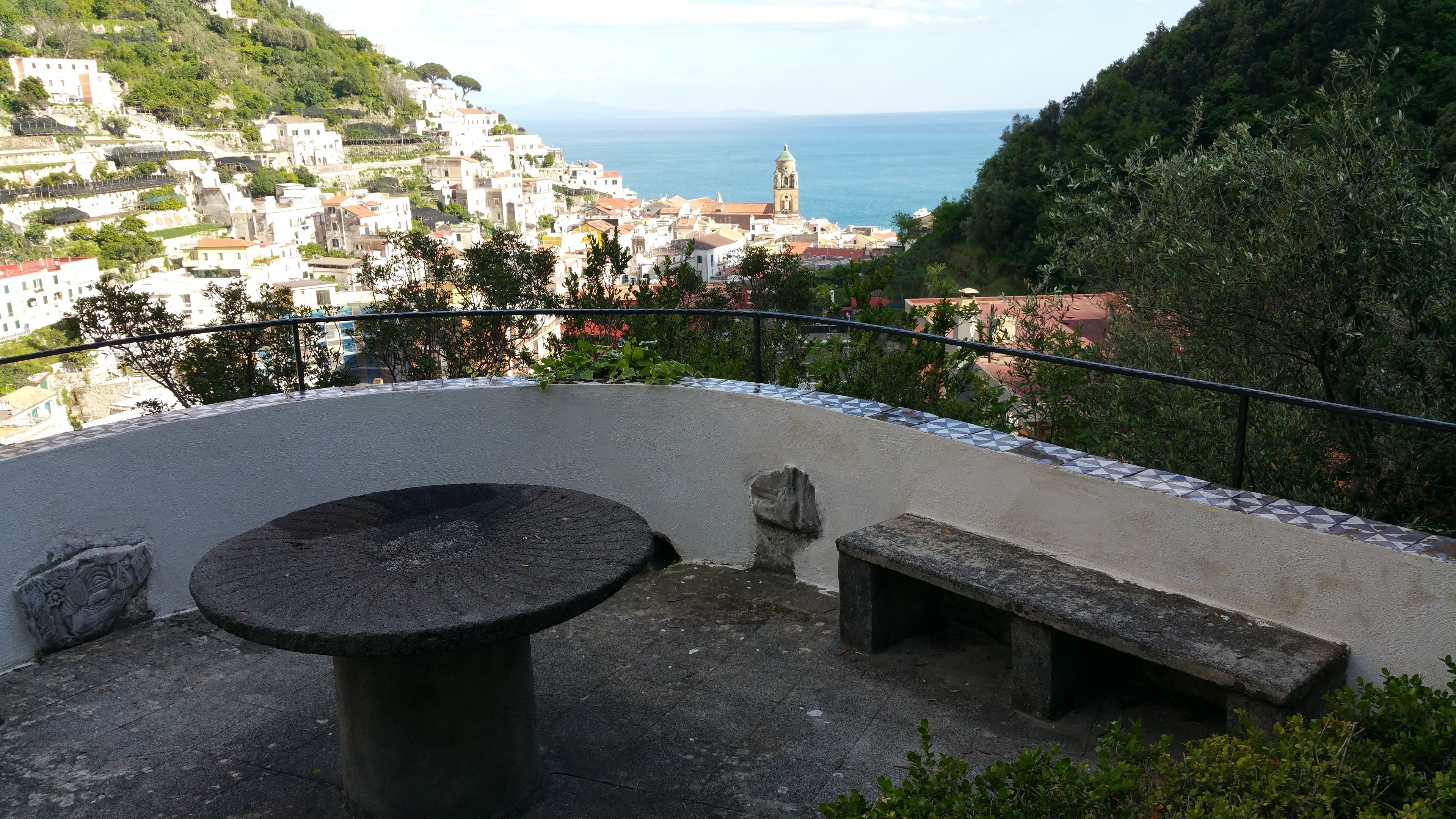 A terrace with a round table and benches, overlooking a coastal town with buildings and a church tower.