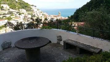 A terrace with a round table and benches, overlooking a coastal town with buildings and a church tower.