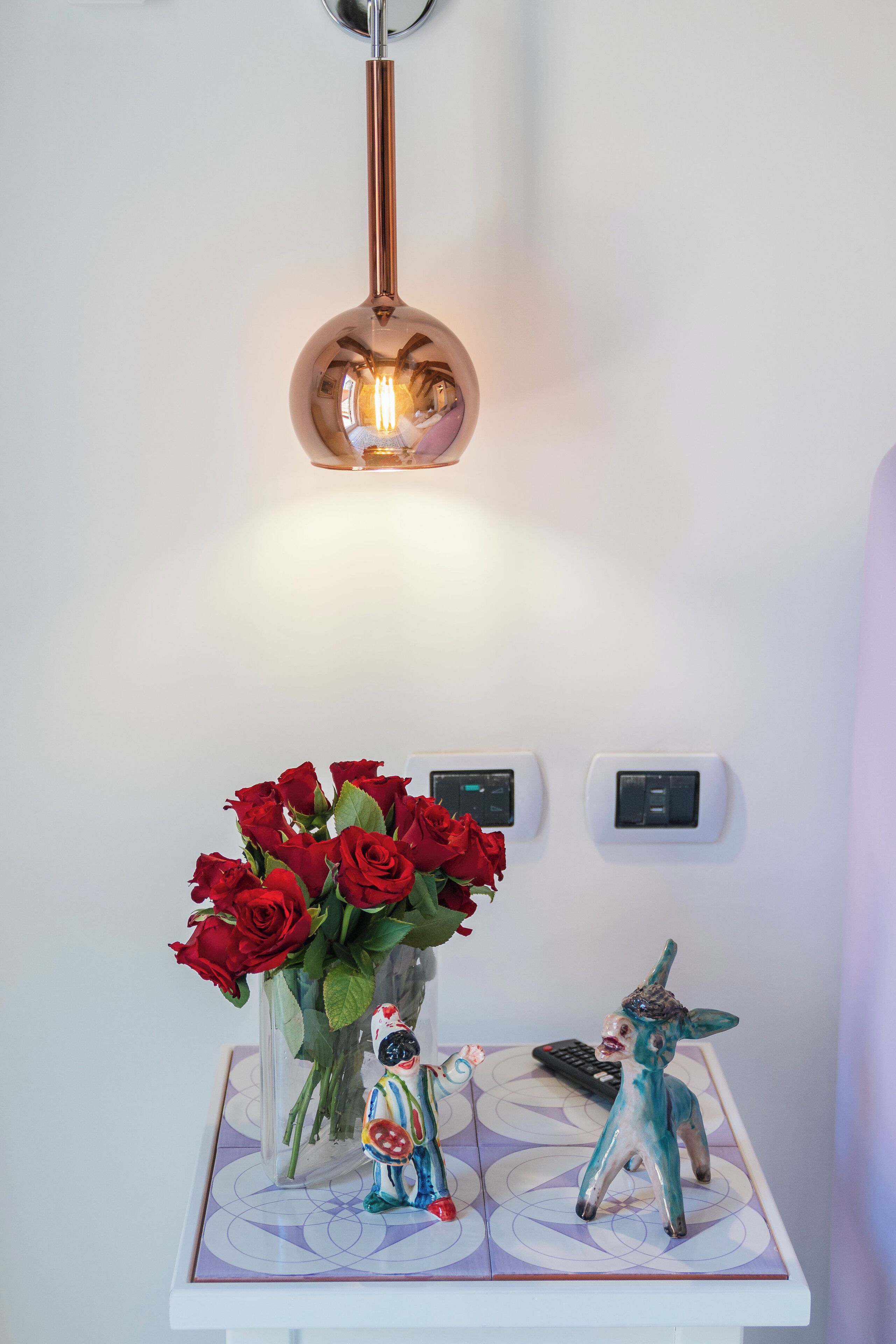 Een wandlamp, een vaas met rode rozen en twee beeldjes op een decoratieve tafel.