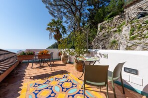 Une terrasse extérieure avec un sol carrelé, une table entourée de chaises, des plantes en pot et une vue sur la mer.