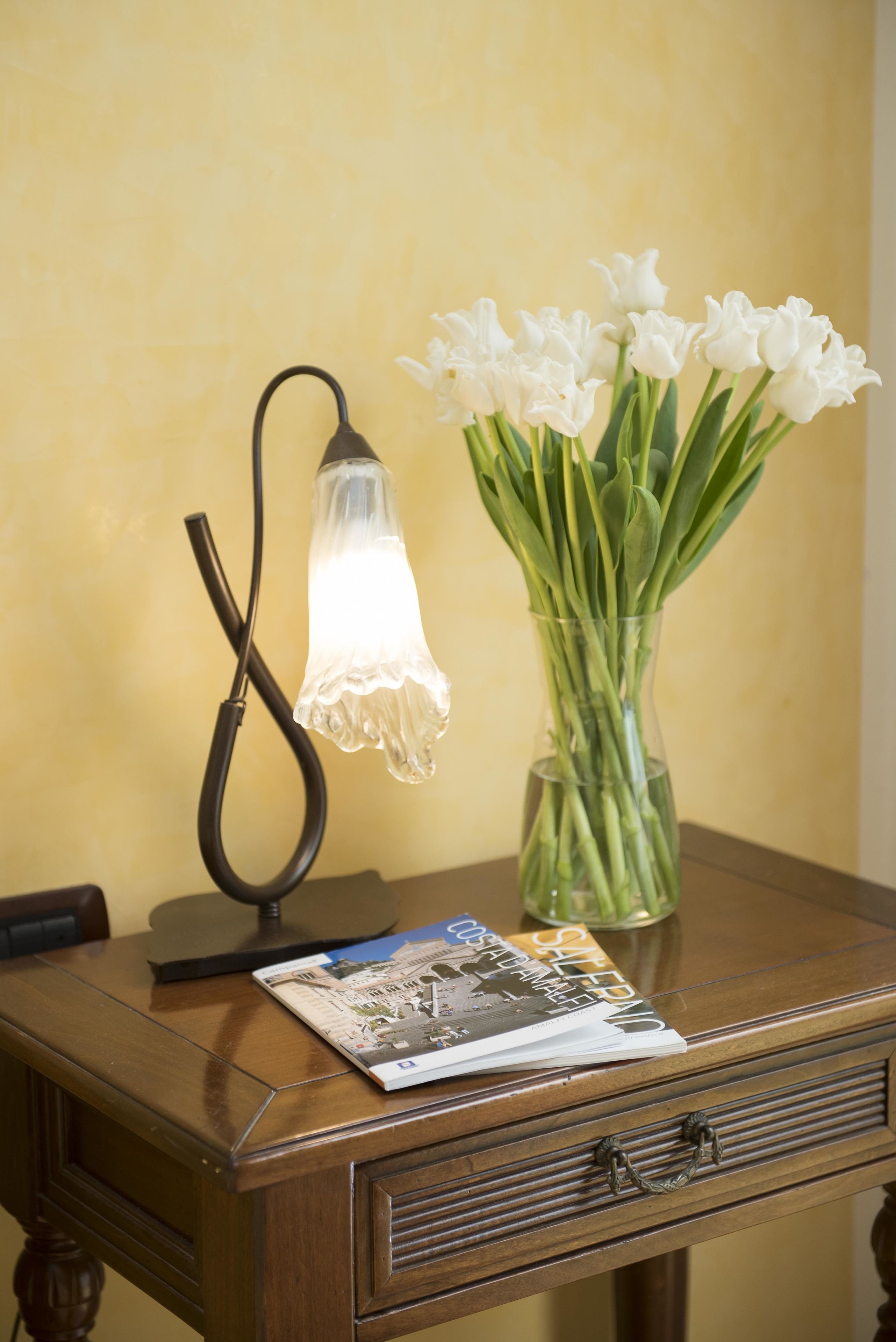Una mesa de madera con una lámpara, un florero con flores blancas y una revista.