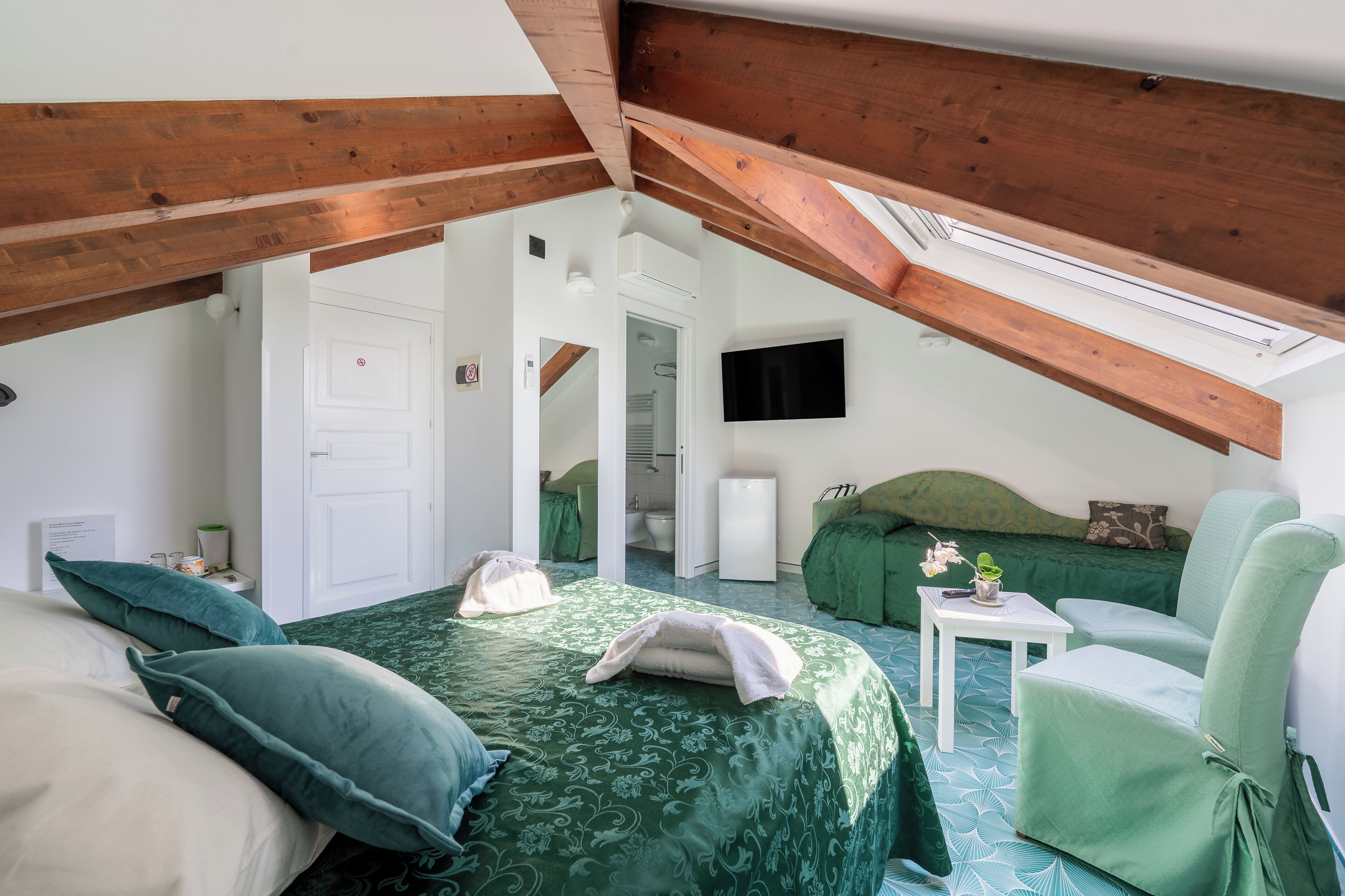 Une chambre avec un plafond en pente, un lit avec une literie verte, un fauteuil vert, une petite table blanche et un téléviseur fixé au mur.