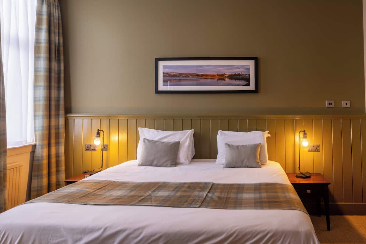A hotel room with a bed, two bedside lamps, a framed picture on the wall, and a window with curtains.