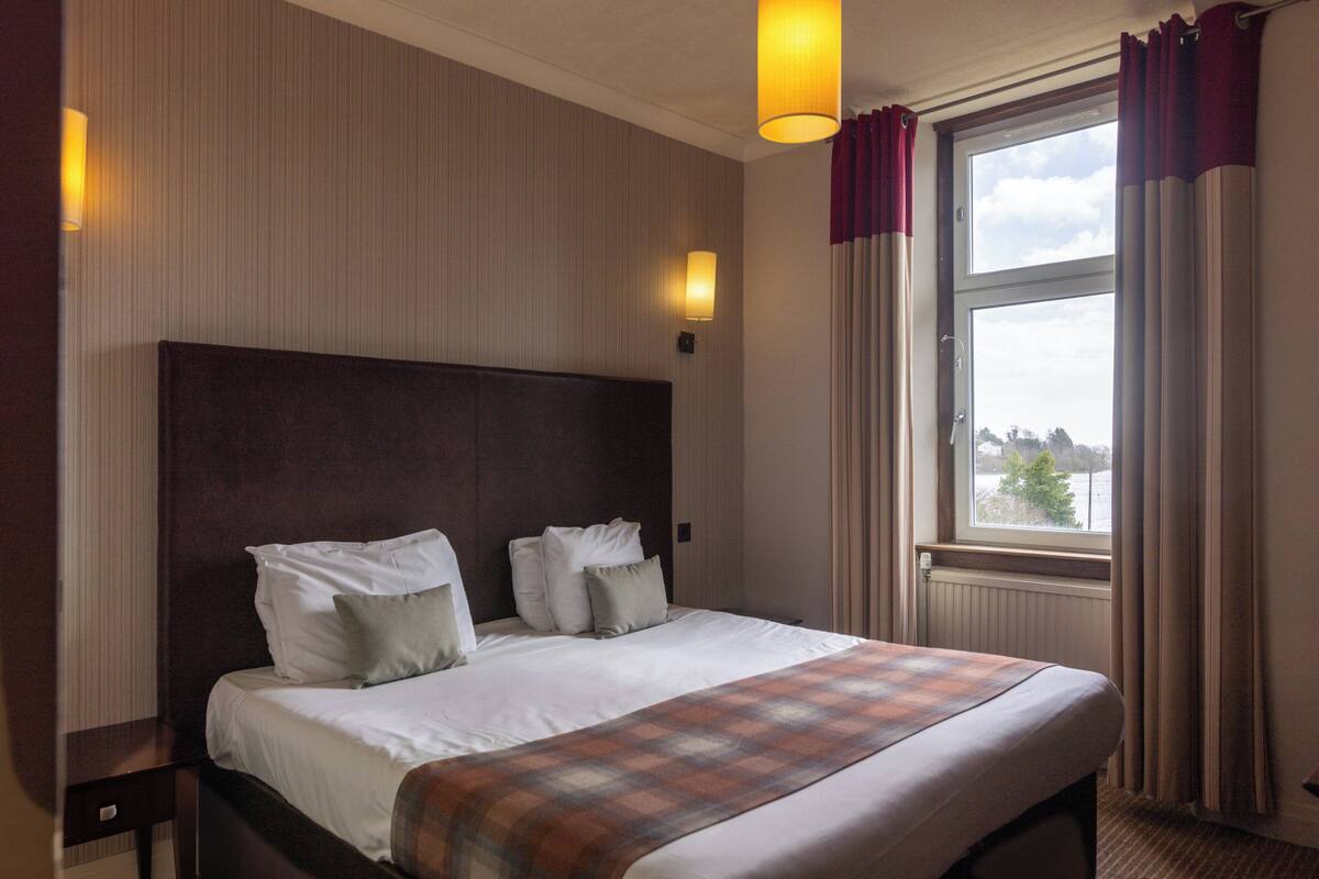 A hotel room with a large bed, two bedside tables, a window with curtains, and wall-mounted lights.