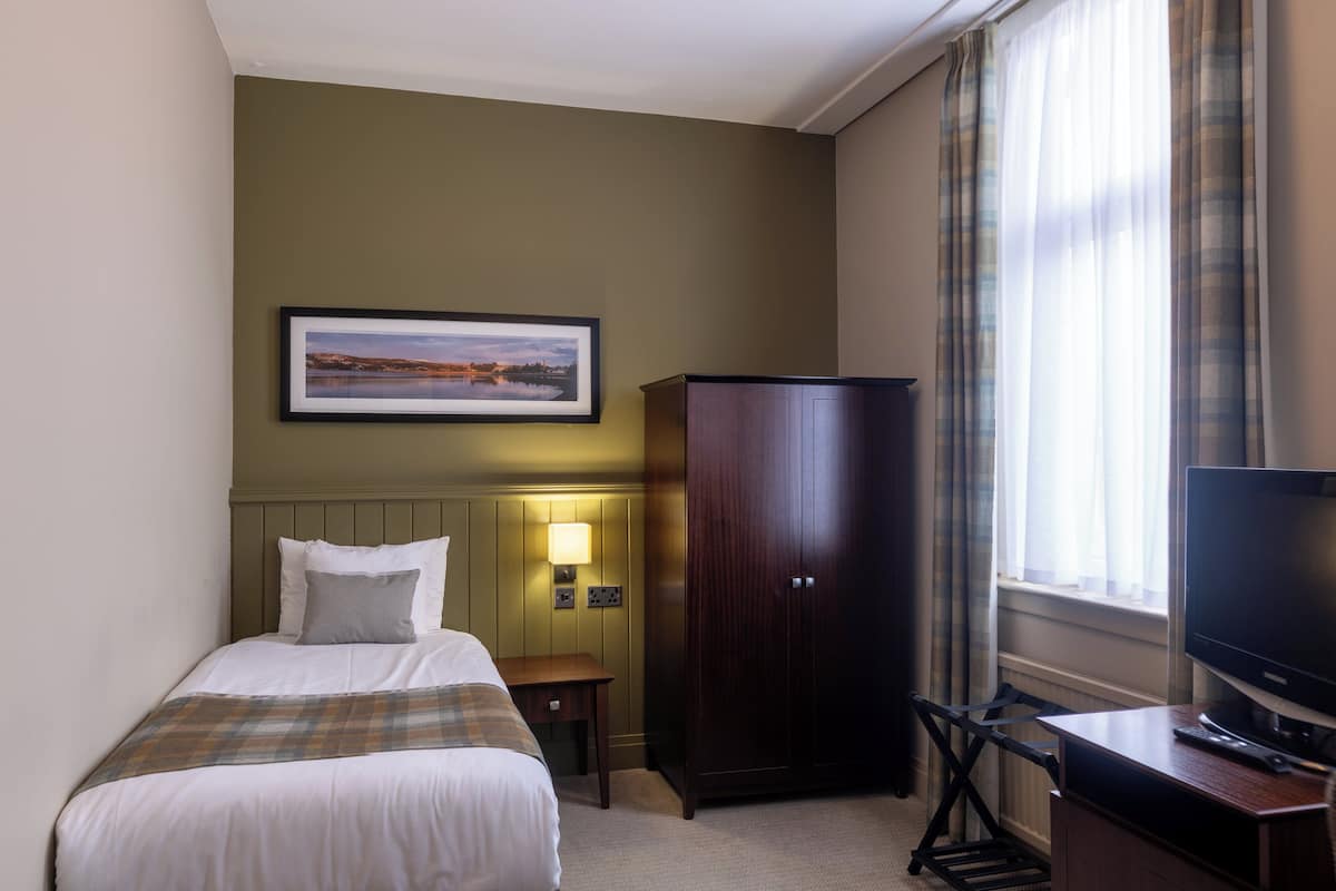 A hotel room with a bed, a desk with a television, a wardrobe, a bedside table with a lamp, and a framed picture on the wall.