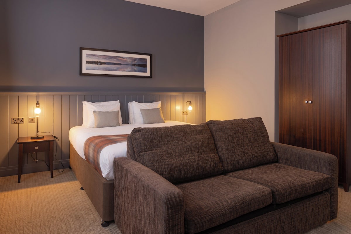 A bedroom with a bed, a brown sofa, a wooden wardrobe, and a framed picture on the wall.