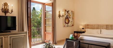 A hotel room with a bed, a TV, a dining table with a book and flowers, a painting, and a view of greenery outside.