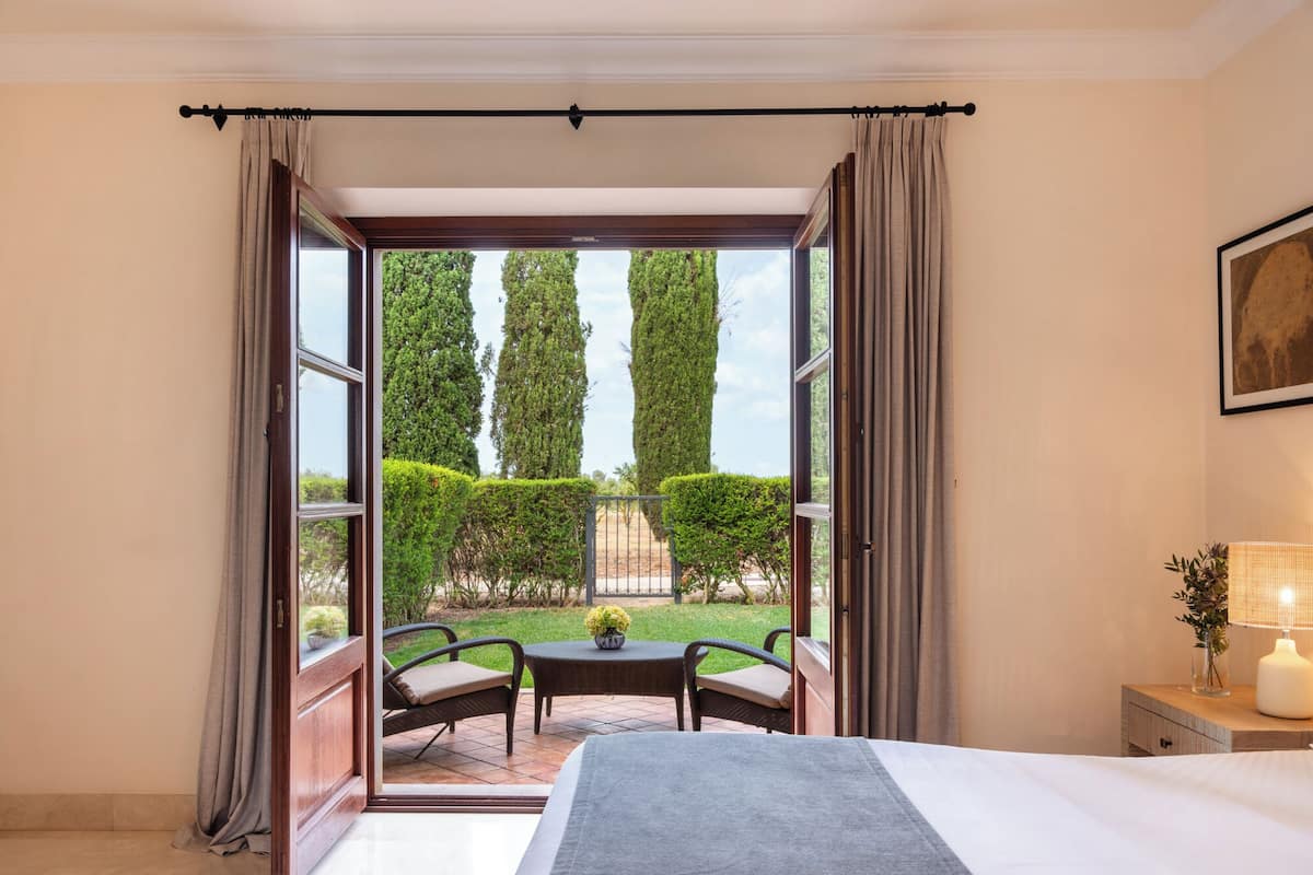 Ein Hotelzimmer mit einem großen Bett, einem kleinen Tisch mit Stühlen und Blick auf einen Garten mit ordentlich geschnittenen Hecken und Bäumen.