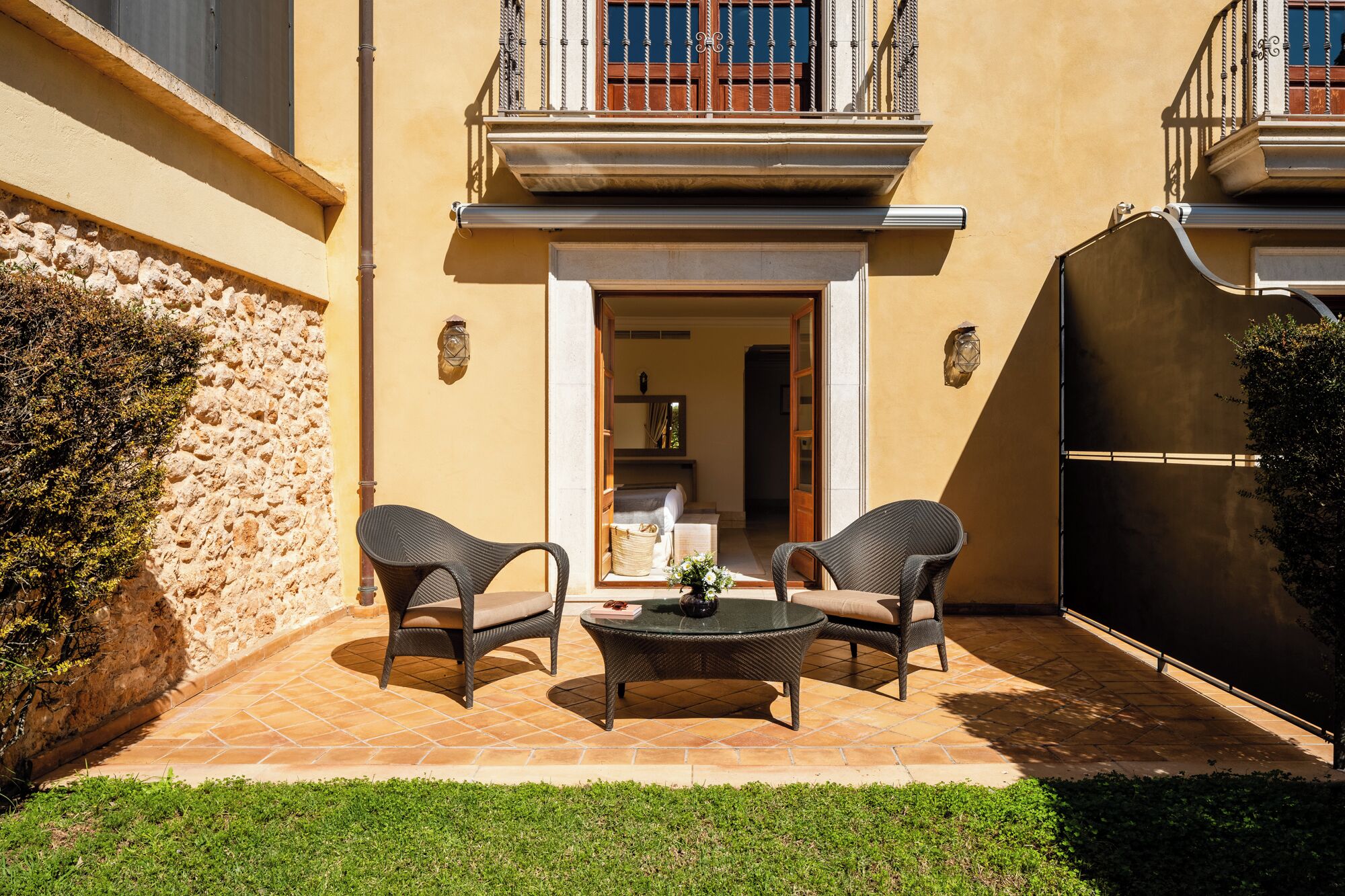Un patio con zona de comedor, dos sillas de mimbre, una mesa redonda y vista al interior del edificio.