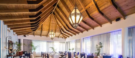 Une salle à manger spacieuse avec de hauts plafonds en bois et des lustres, des tables dressées pour le repas, et de grandes fenêtres ornées de rideaux.