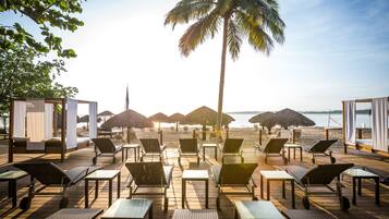 A beachside lounge with sun loungers and tables, palm trees, and umbrellas.