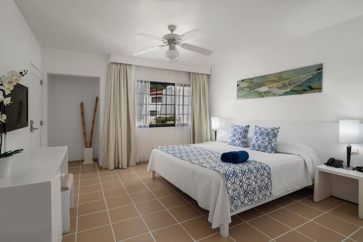 Habitación de hotel con cama, mesitas de noche, ventilador de techo, ventana con cortinas, un cuadro en la pared y una planta en maceta.