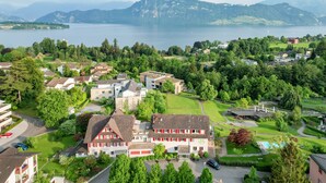 A residential area with a large house featuring a red garage, surrounded by lush greenery and a lake in the background. - Hotel Restaurant Balm Meggen (Meggen)