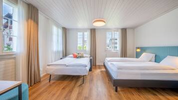 A modern hotel room with two beds, wooden flooring, large windows with curtains, and a blue accent wall.