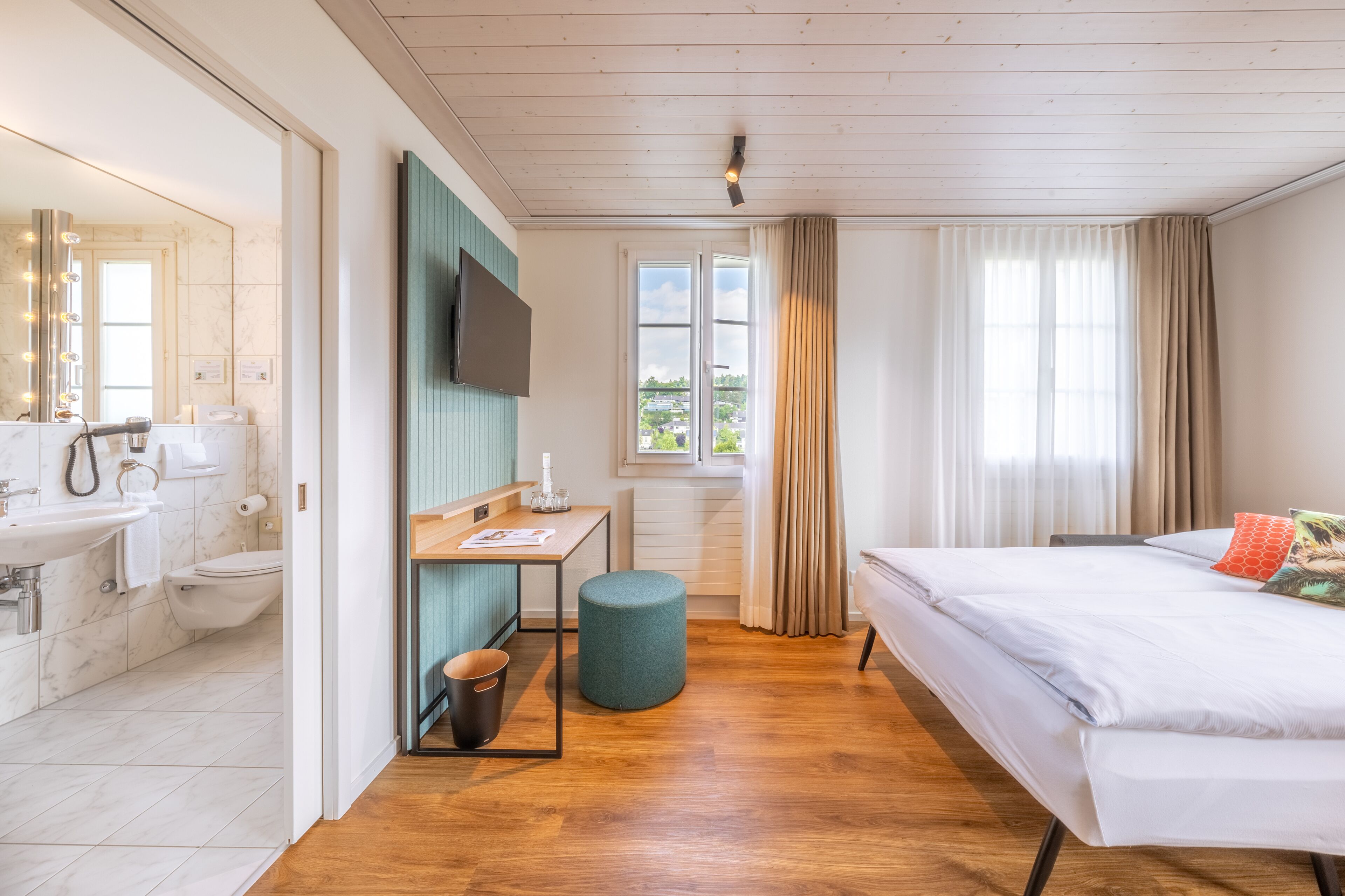 A modern hotel room with a wooden floor, a large bed, a desk with a chair, a television, and a bathroom with a mirror and sink.