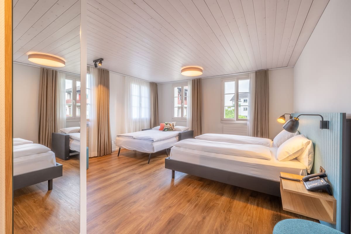 A modern hotel room with three beds, wooden flooring, large windows, and a mirror reflecting the room.