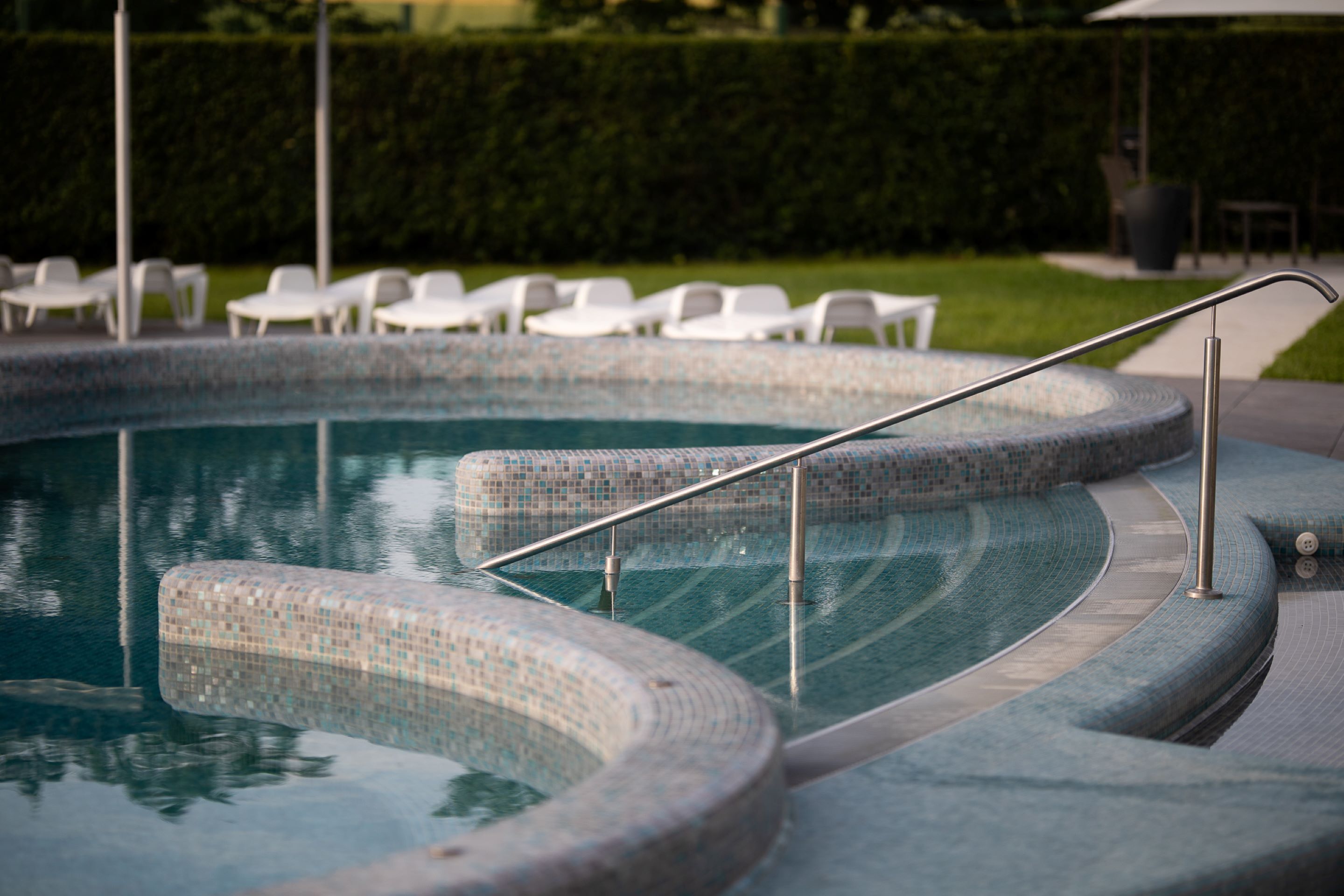 Une piscine ornée de motifs en mosaïque, avec une rampe métallique et, au fond, des chaises longues blanches.