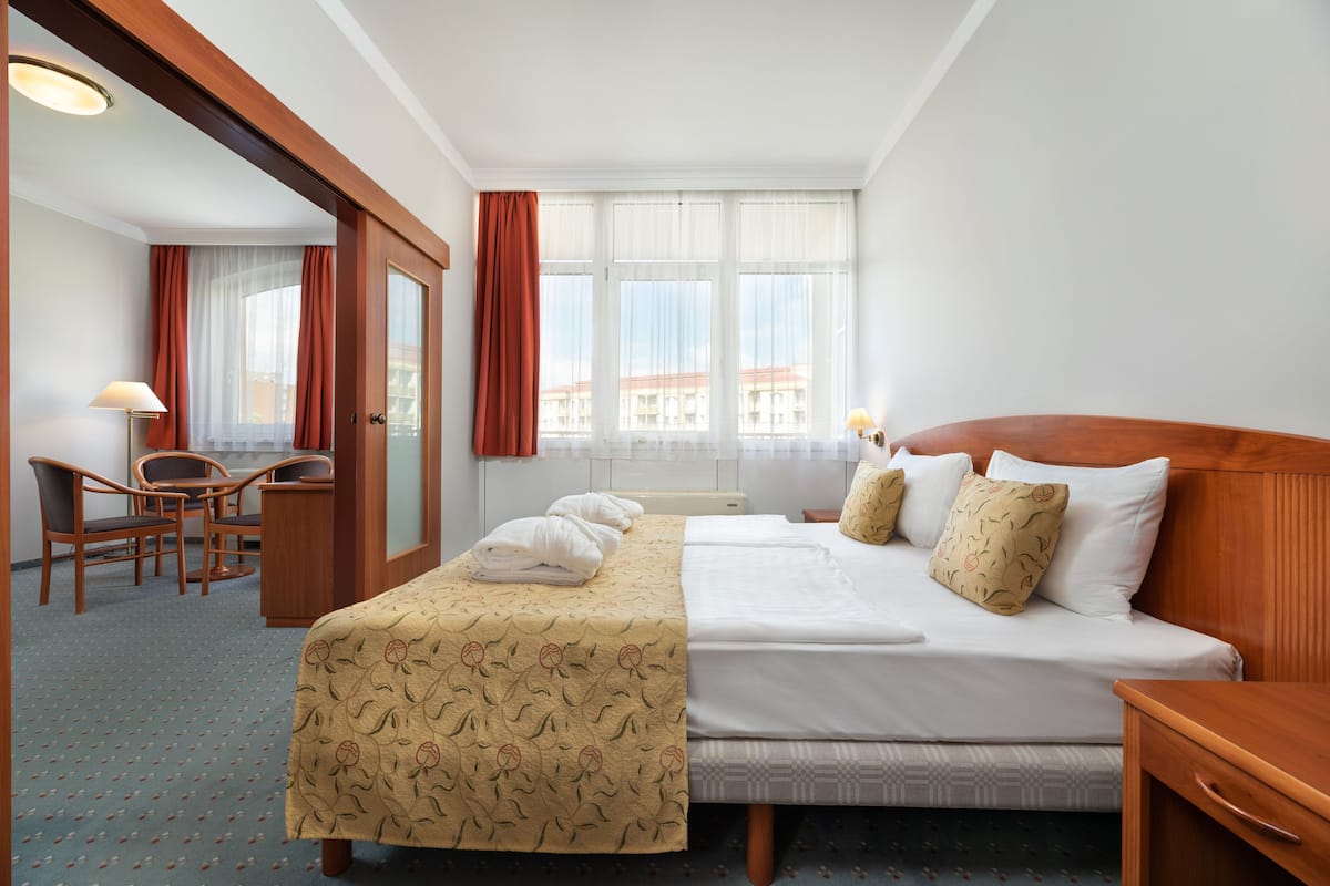 Quarto de hotel com cama, duas almofadas, mesa de cabeceira, escrivaninha com cadeira e janela com cortinas.