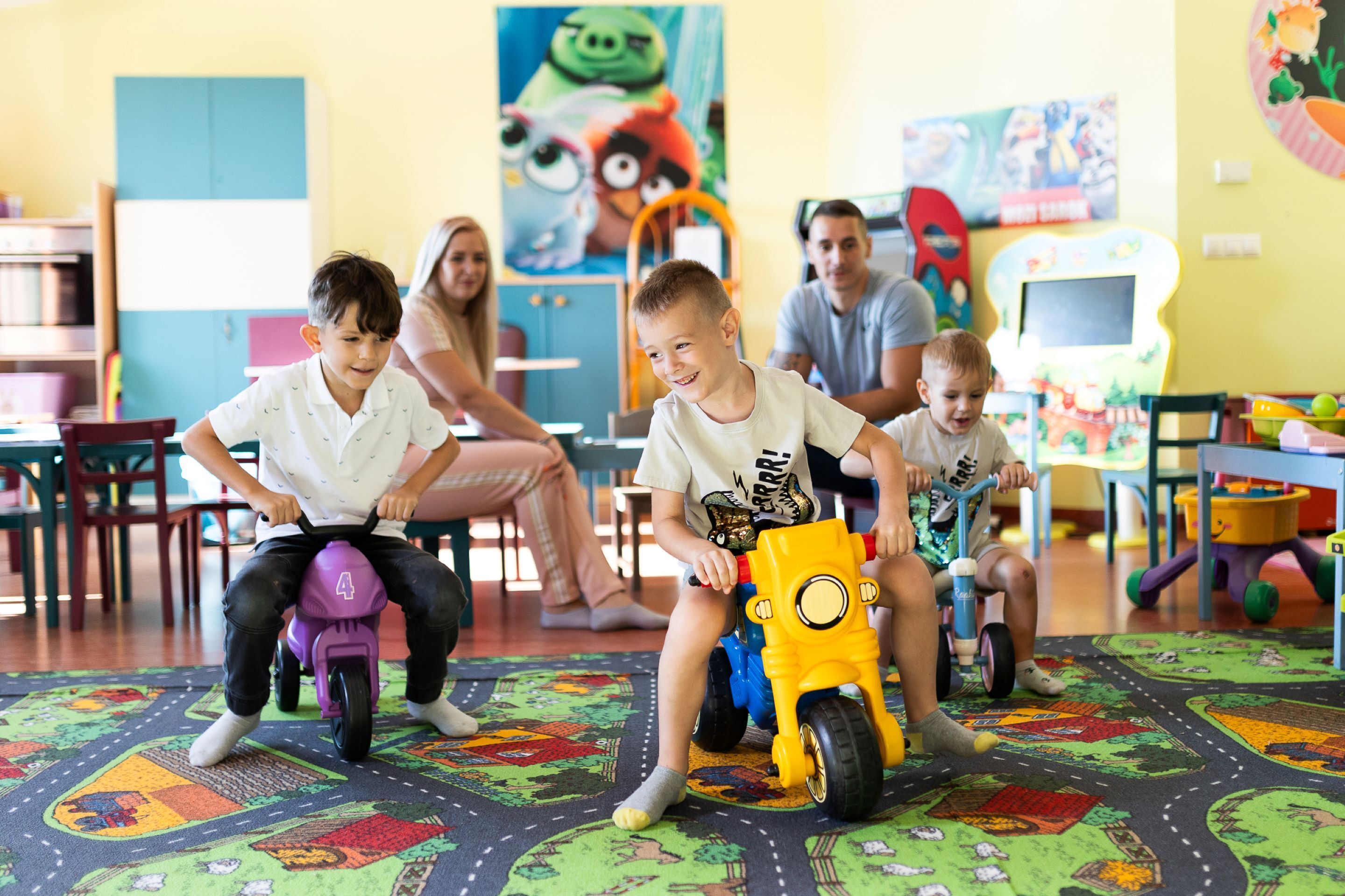 Children riding toy motorcycles in a brightly colored playroom with educational posters on the walls.