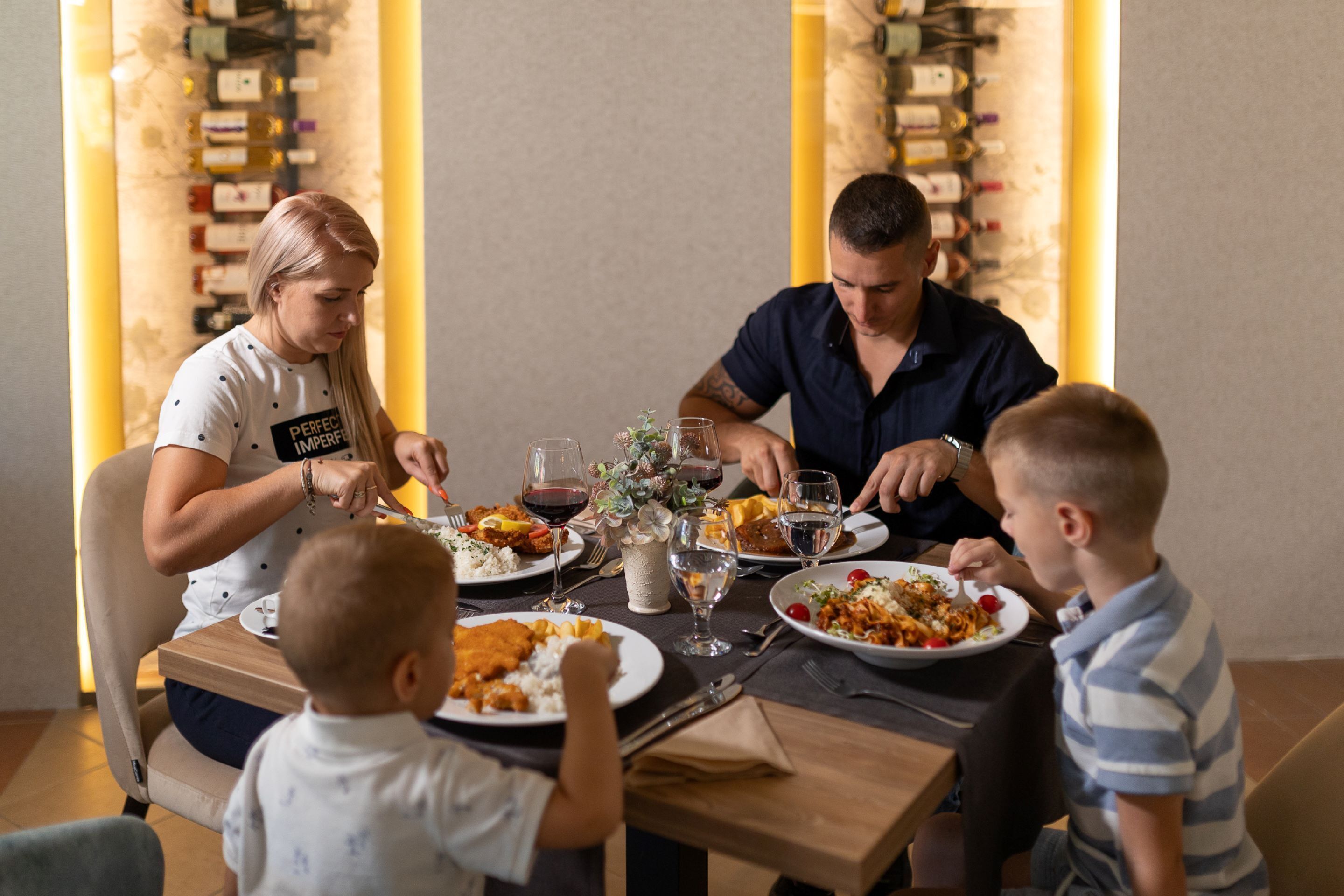 Una familia cenando en un restaurante, con una estantería llena de vino al fondo.