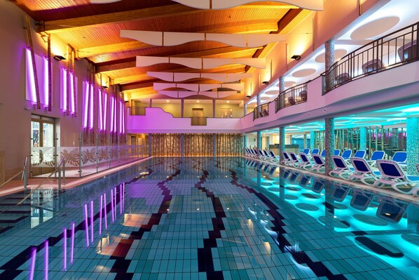 A spacious indoor swimming pool with a tiled floor and blue lighting, surrounded by lounge chairs and a wooden ceiling with unique patterns. - Hotel Karos Spa (Zalakaros)