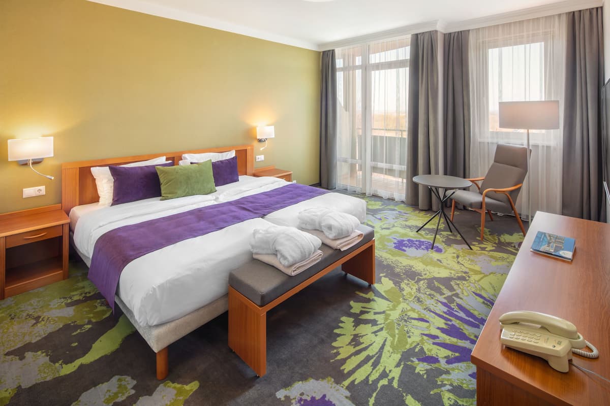 Quarto de hotel com cama, mesas de cabeceira, escrivaninha com telefone, cadeira e janela com cortinas.