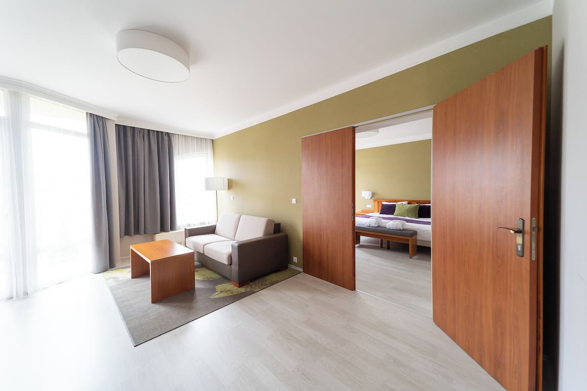 Quarto de hotel moderno com sofá, uma pequena mesa de madeira, cama e uma janela grande com cortinas.