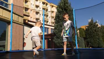 Two children playing on a trampoline in front of a building with multiple balconies.