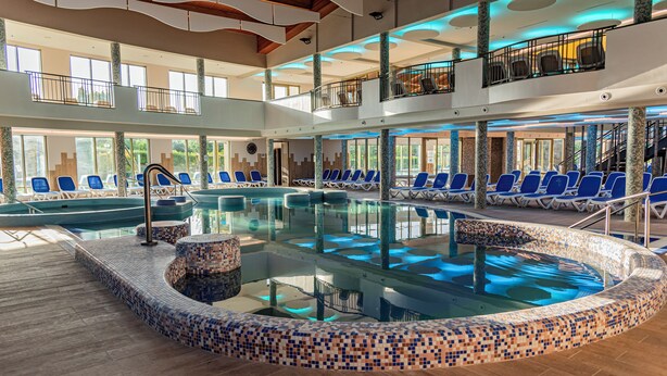 Una gran piscina interior con sillas azules alrededor, un balcón en el segundo piso con sillas y un suelo de baldosas.