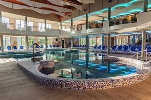 Una gran piscina interior con sillas azules alrededor, un balcón en el segundo piso con sillas y un suelo de baldosas.