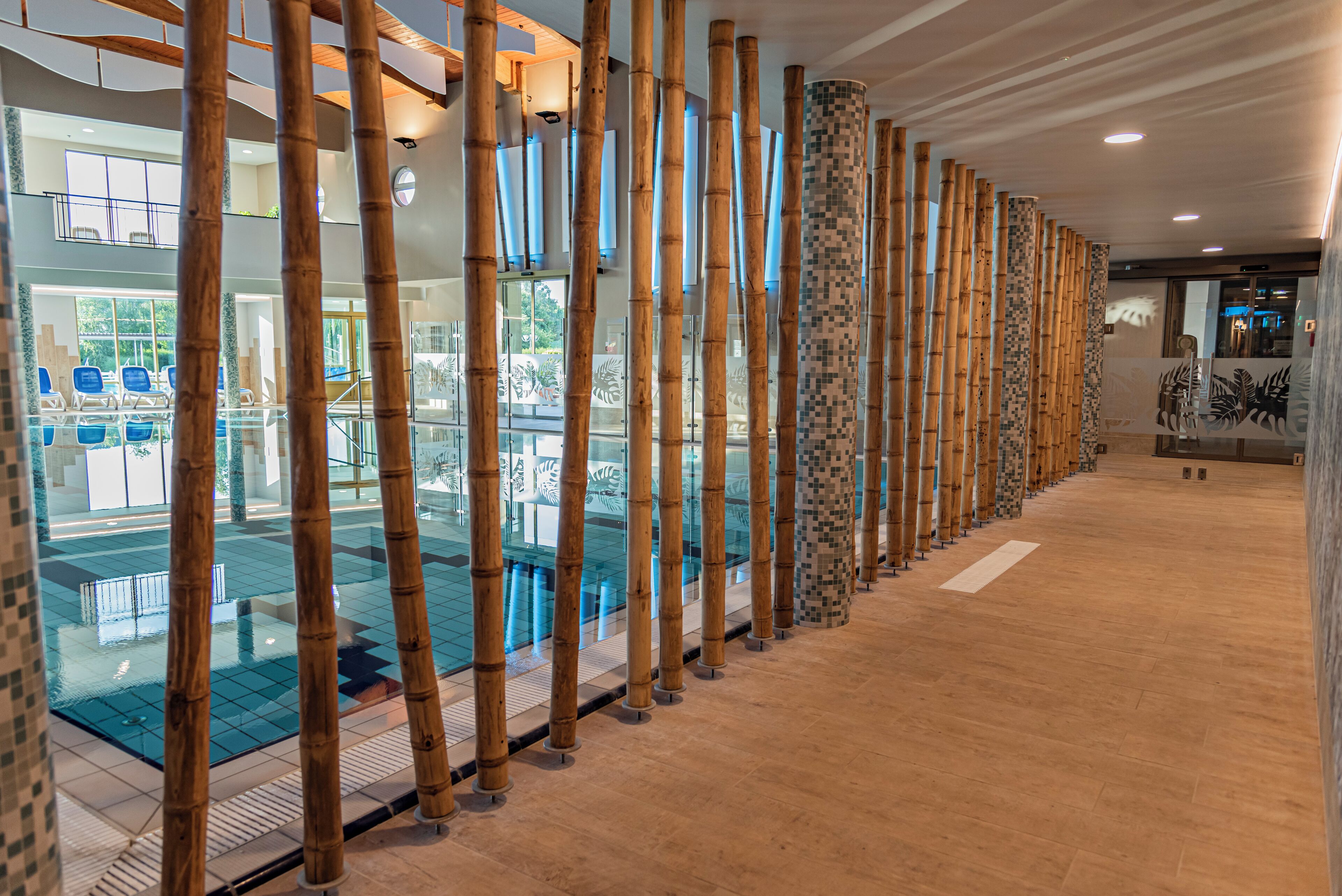 Un espace piscine intérieur moderne, agrémenté de colonnes en bambou, de rampes en verre et d’un sol carrelé.