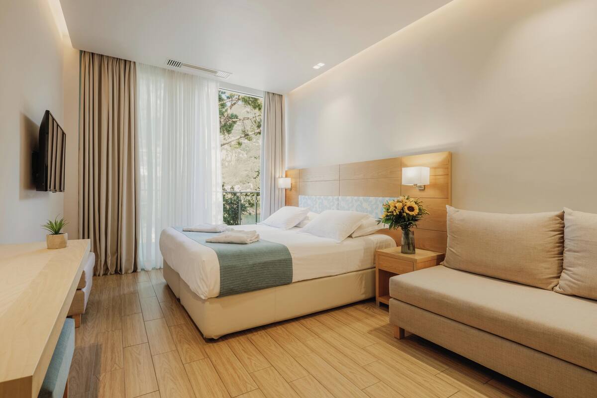 A modern hotel room with a bed, a sofa, a TV, a bedside table with a lamp, and a window with curtains.