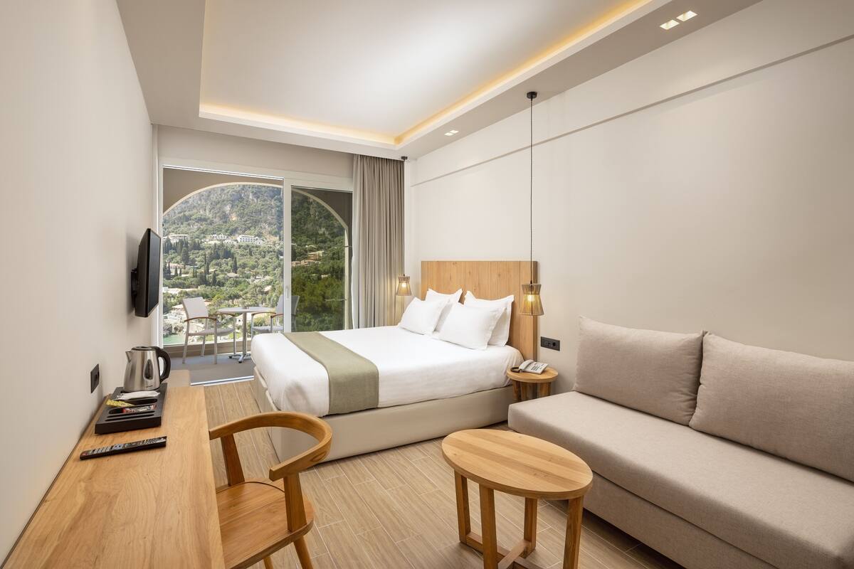 Une chambre d’hôtel moderne équipée d’un lit, d’un canapé, d’un bureau avec une chaise, d’une télévision et offrant une vue sur un paysage montagneux.