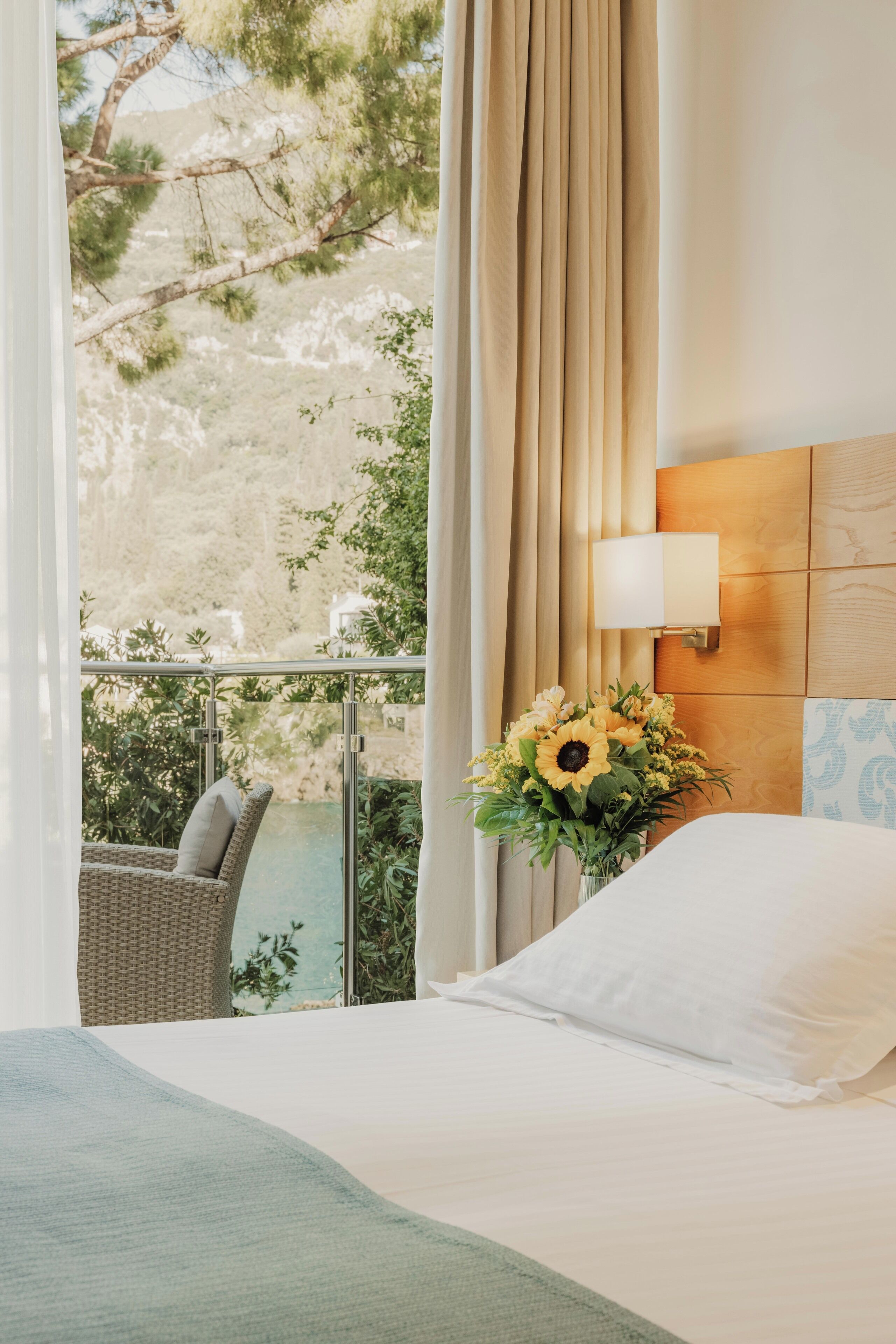 A hotel room with a bed, a view of a mountainous landscape, a wicker chair, and a vase with flowers.