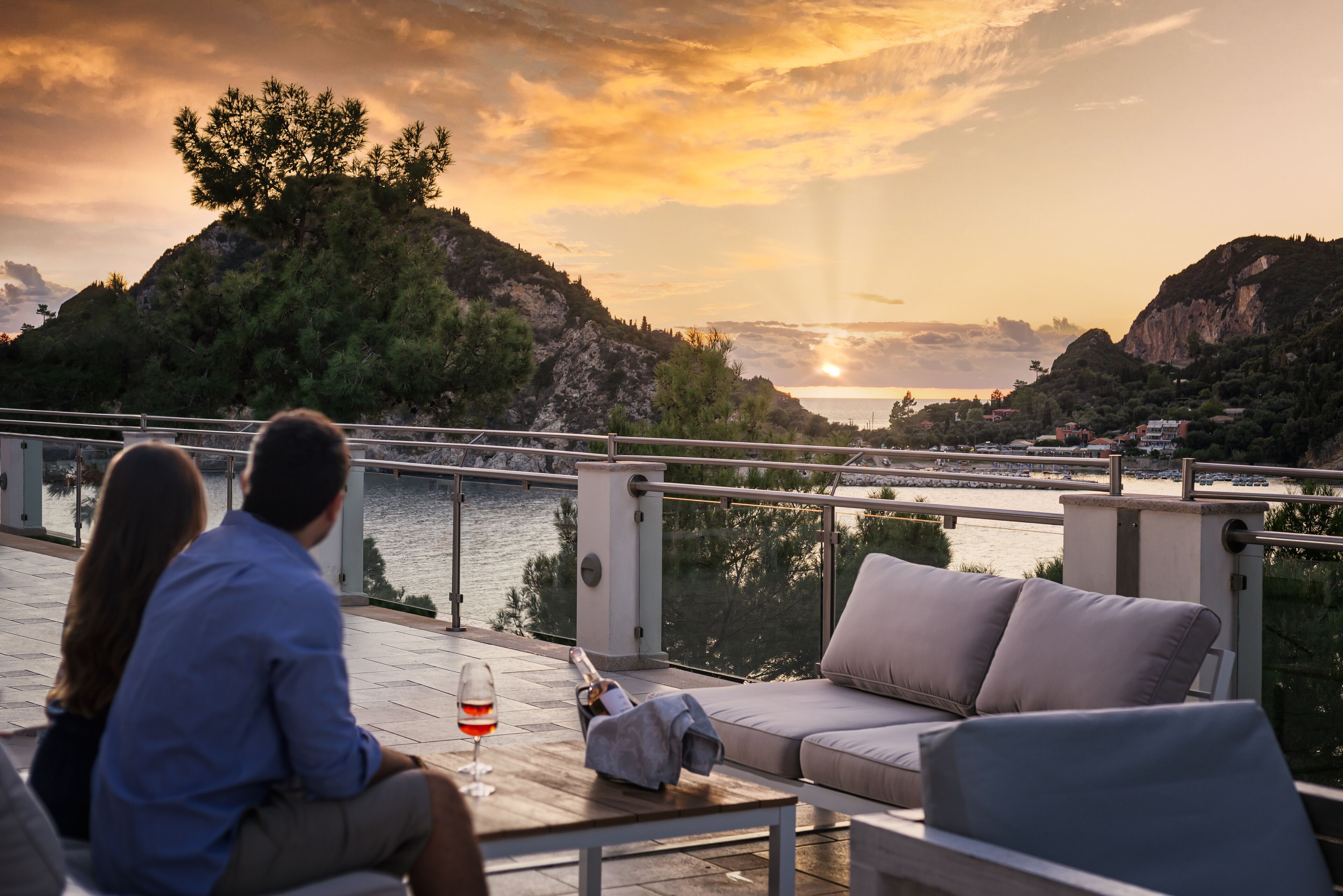 A couple seated on a balcony overlooking a scenic view of a body of water and hills during sunset.