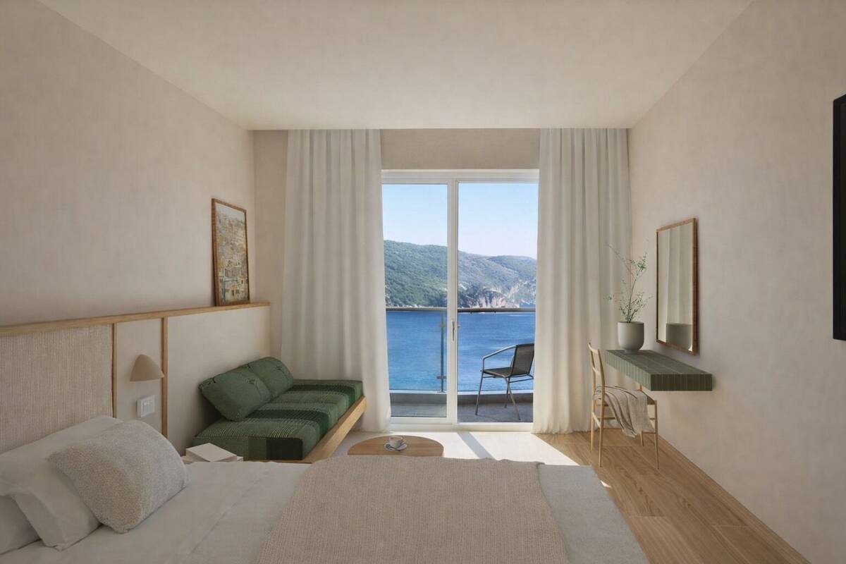 Une chambre avec un lit, un canapé vert, une chaise, une petite table, un miroir et une vue sur la mer et les collines.