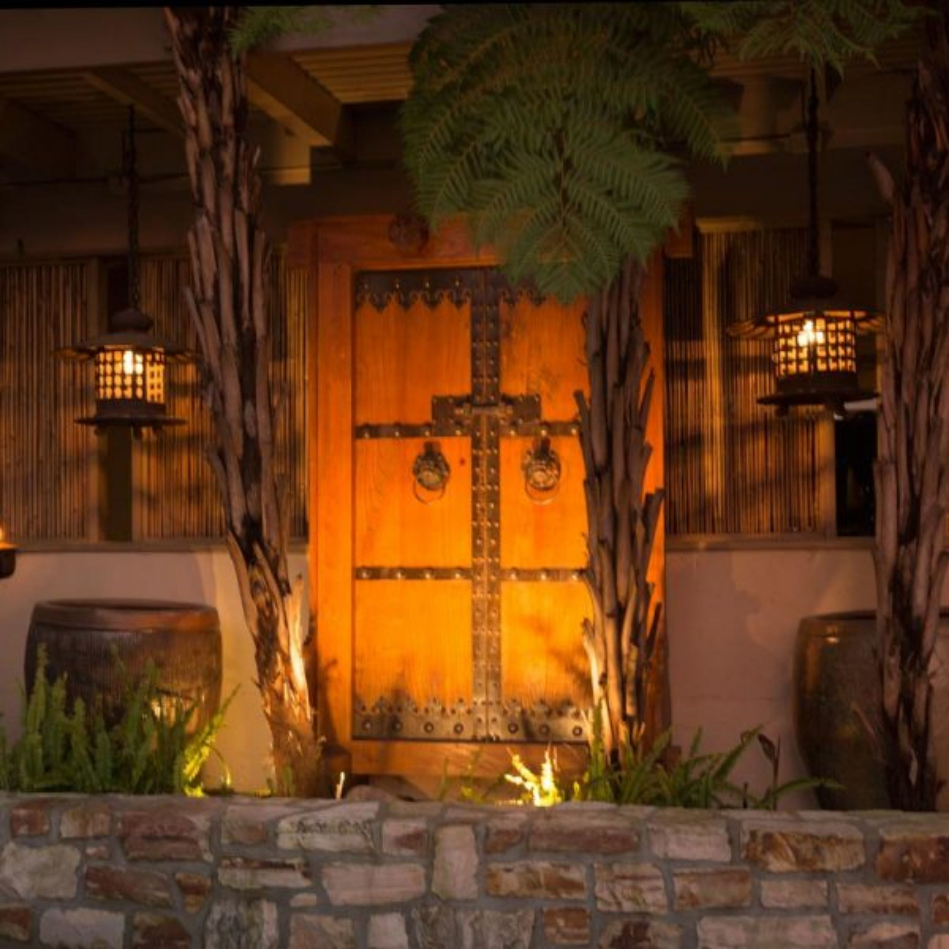 A wooden door with a cross, flanked by lanterns and plants, leading to a stone wall.
