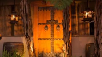 A wooden door with a cross, flanked by lanterns and plants, leading to a stone wall.