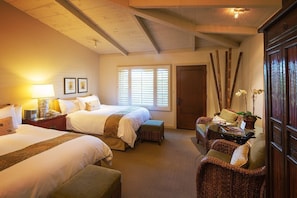A hotel room with two beds, a sitting area with wicker furniture, a wooden door, and a window with blinds.