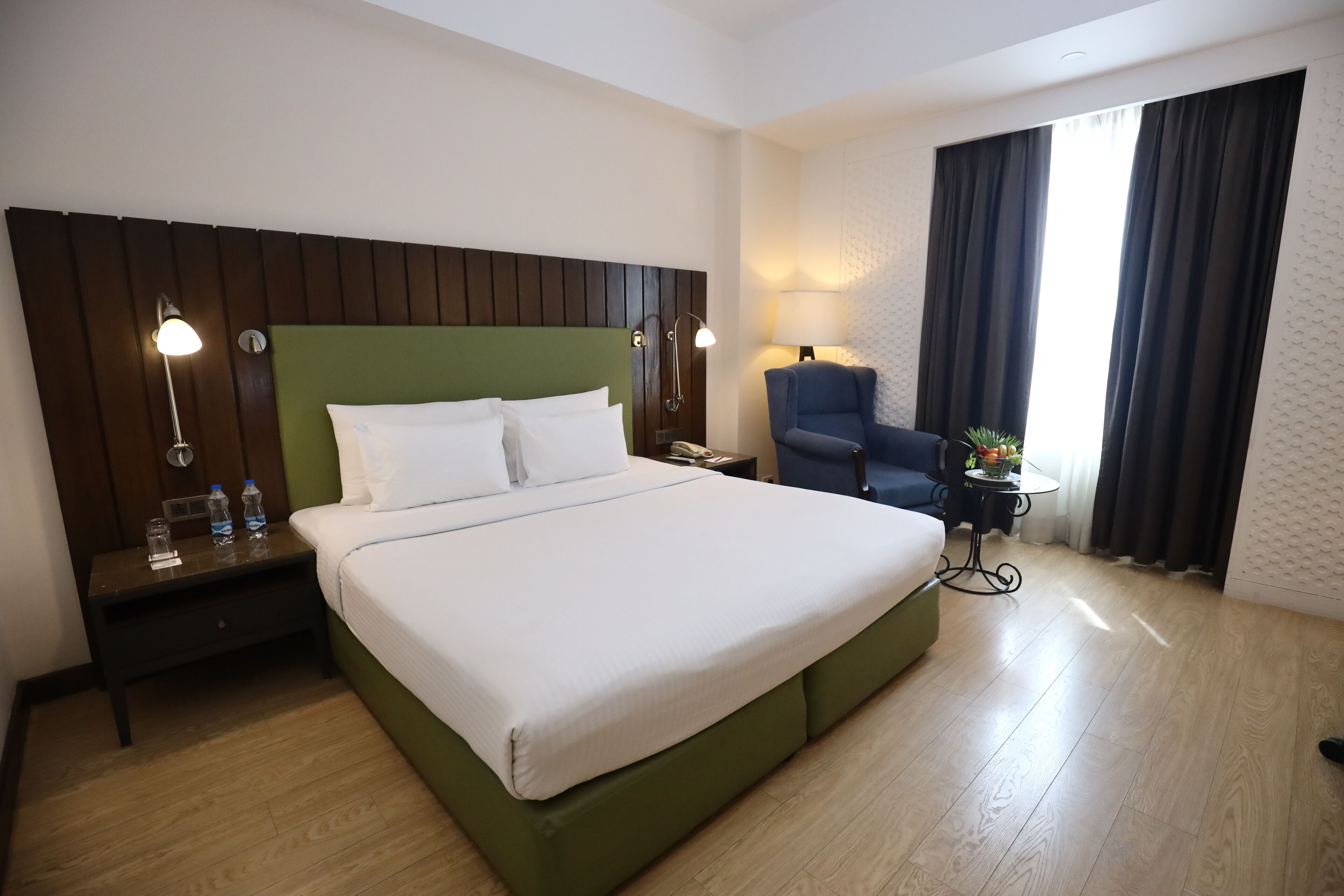 A hotel room with a large bed, a nightstand with a lamp, a chair, and a window with curtains.