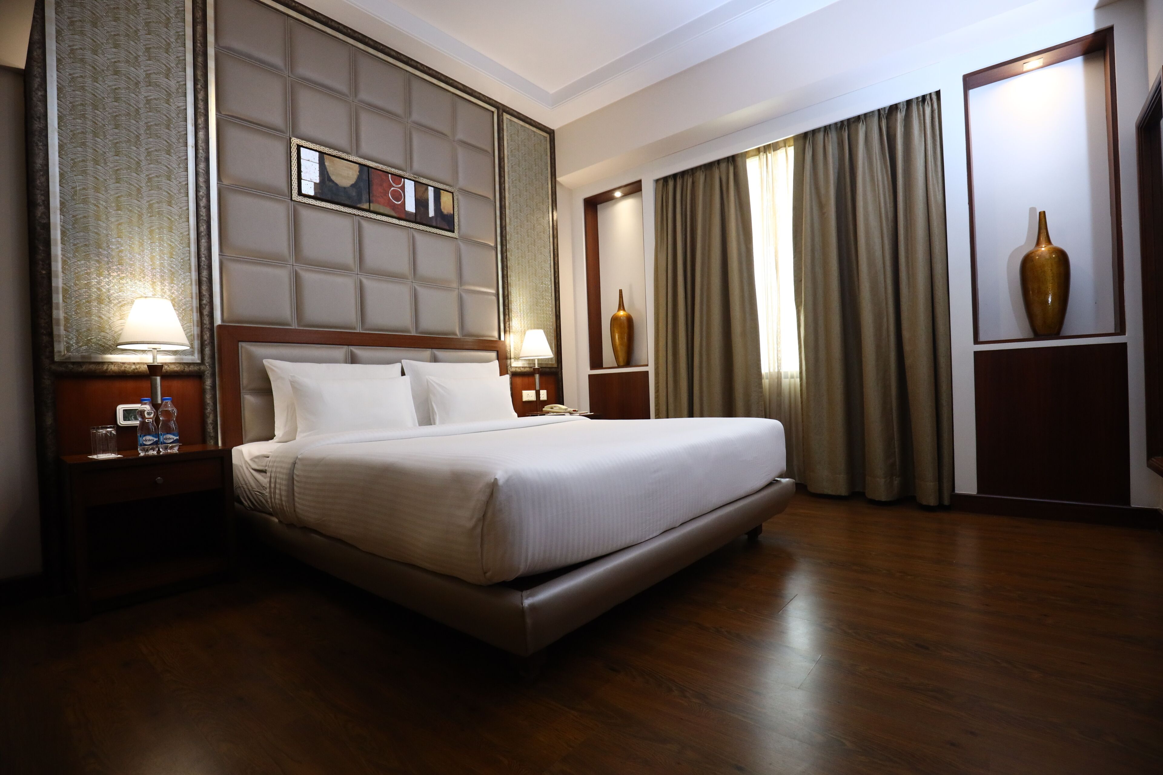 A hotel room with a large bed, wooden floor, a headboard with a decorative panel, a nightstand with a lamp, and a vase on the wall.