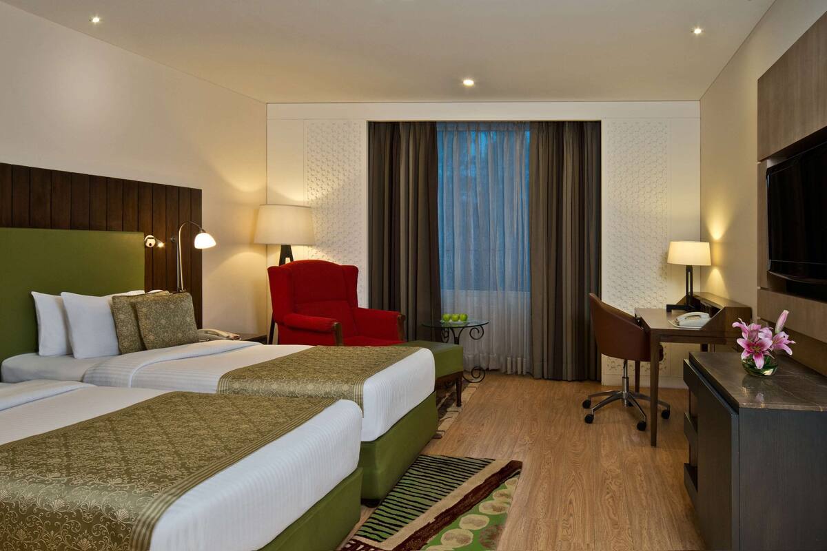 Une chambre d’hôtel avec deux lits, un bureau, une chaise rouge, une télévision et une fenêtre avec des rideaux.