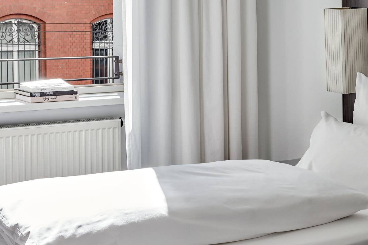 A hotel room with a bed, a window with a view of a brick building, and a radiator.