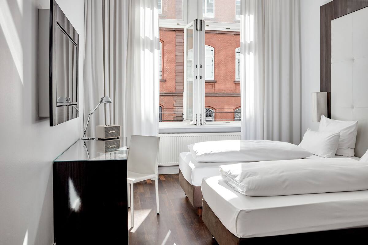 A modern hotel room with two beds, a desk with a lamp, and a view of a red brick building through the window.