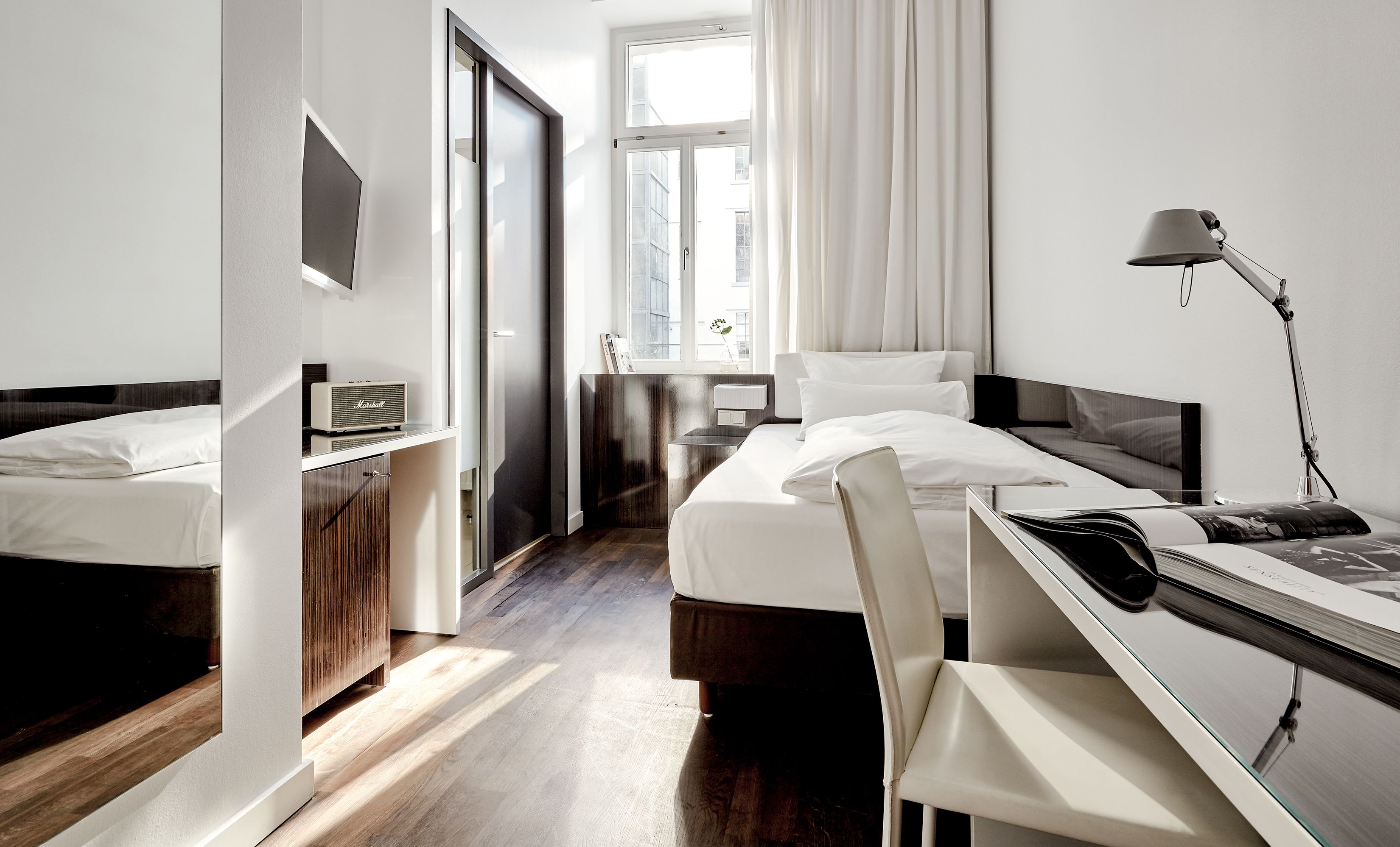 Ein Hotelzimmer mit zwei Betten, einem Schreibtisch, einem Stuhl, einer Lampe, einem Radio und einem Fenster mit Vorhängen.