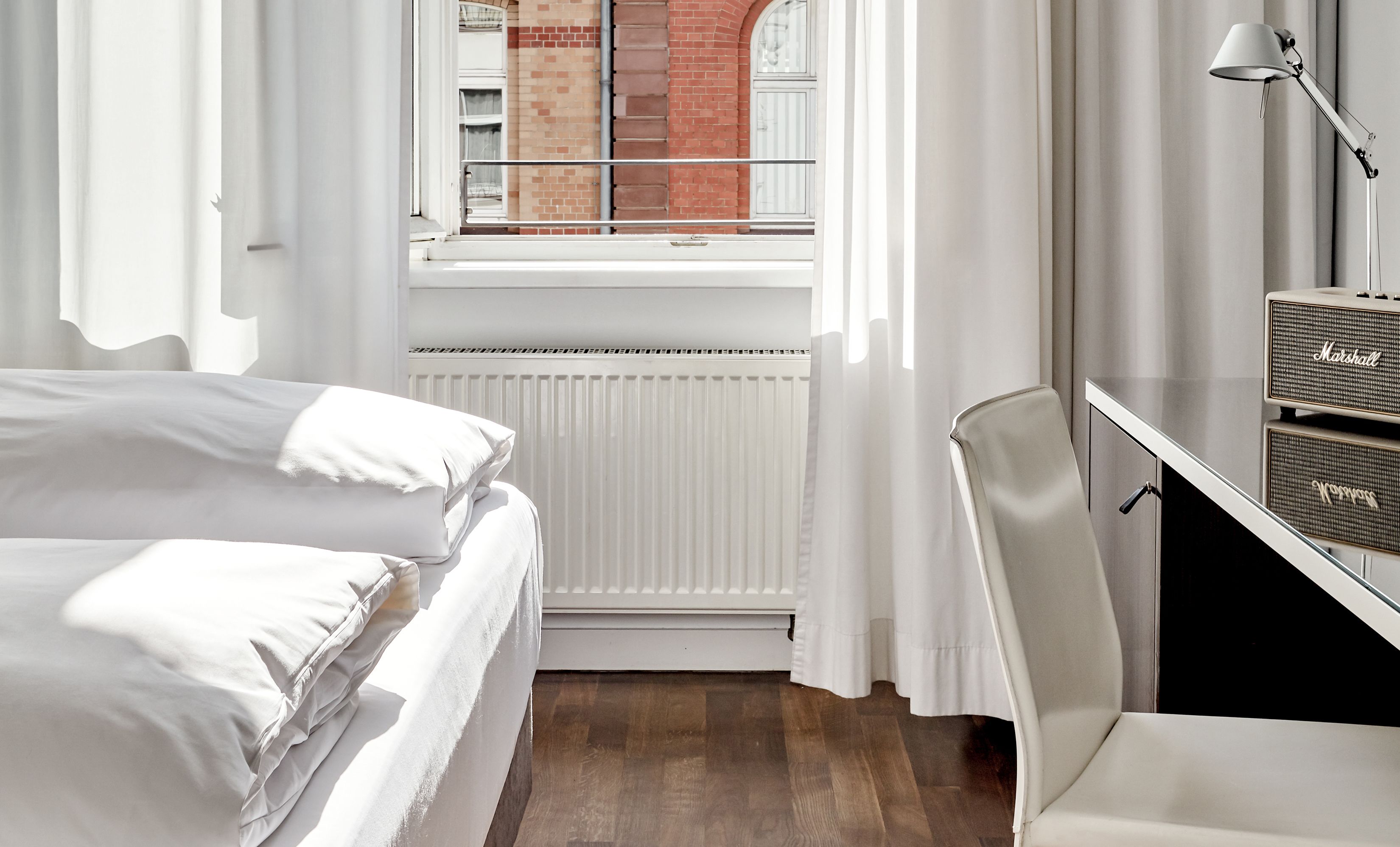 A hotel room with a bed, a desk with a lamp, a radiator, and a window with curtains.