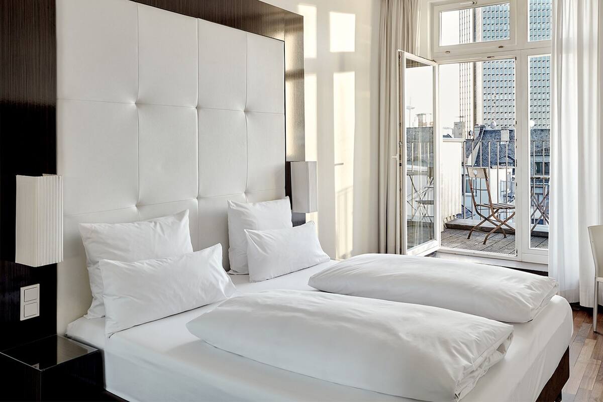 A modern hotel room with a large bed, a desk, and a view of the cityscape through large windows.