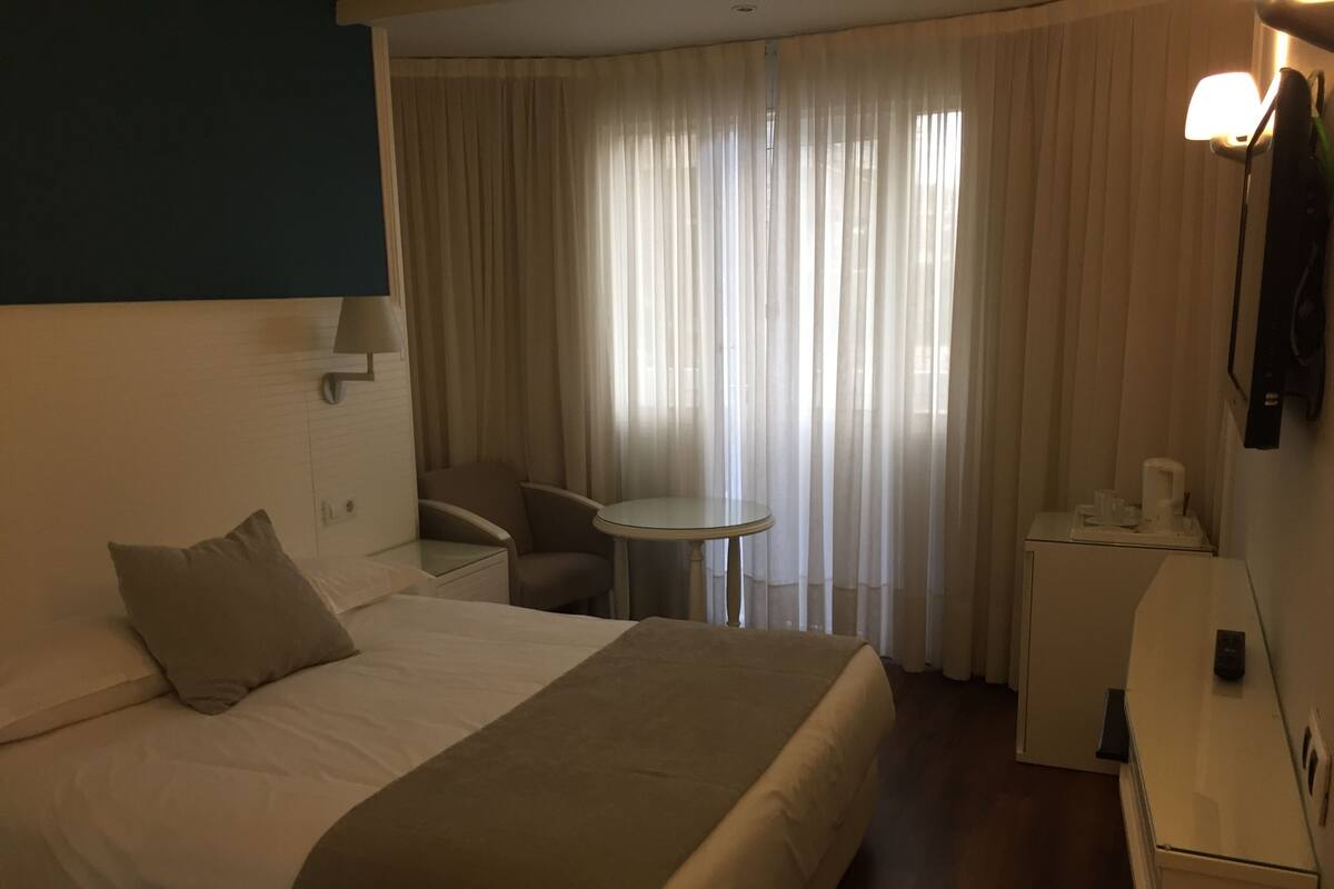 Habitación de hotel con cama, escritorio, silla, mesita y ventana con cortinas.